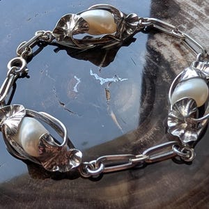 May include: A silver bracelet featuring three large, creamy white pearls encased in ornate, leaf-like settings. The bracelet is connected by a series of small, linked silver chains. The design is intricate and elegant, with a vintage aesthetic.