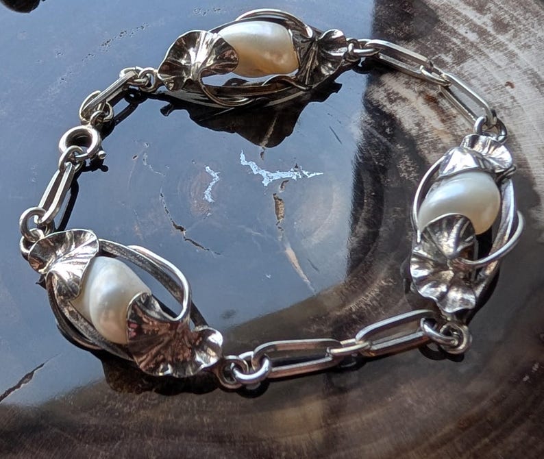May include: A silver bracelet featuring three large, creamy white pearls encased in ornate, leaf-like settings. The bracelet is connected by a series of small, linked silver chains. The design is intricate and elegant, with a vintage aesthetic.