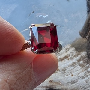 May include: A silver ring featuring a large, rectangular, faceted, deep red gemstone. The ring's band has an ornate, textured design. The gemstone is held in place by four prongs.