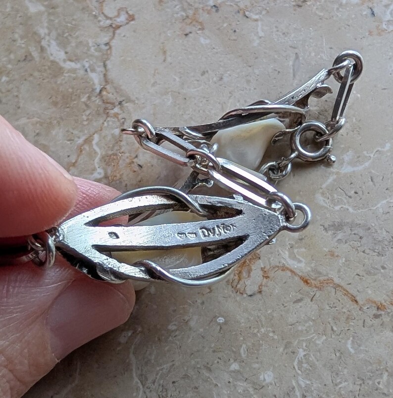 May include: Close-up of a vintage silver necklace with a pearl-like centerpiece. The necklace features an intricate design with a clasp and connecting links. The piece is held by a hand, showcasing the details of the jewelry.