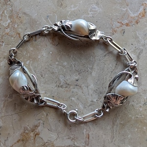 May include: A silver bracelet featuring three large, creamy white pearls. The pearls are set in ornate, leaf-shaped settings connected by a chain of silver links. The bracelet has a secure clasp.