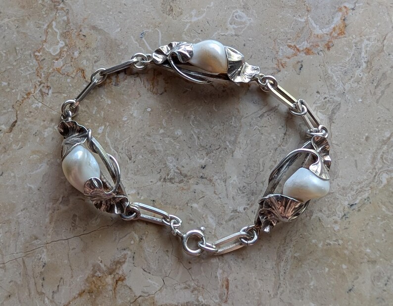 May include: A silver bracelet featuring three large, creamy white pearls. The pearls are set in ornate, leaf-shaped settings connected by a chain of silver links. The bracelet has a secure clasp.