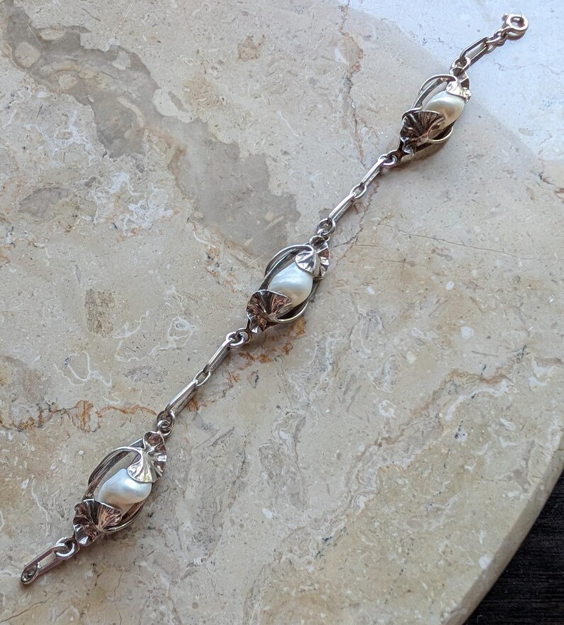 May include: A silver bracelet featuring three large, lustrous pearls encased in decorative settings. The bracelet has a delicate chain design with intricate floral accents, creating an elegant and timeless piece of jewelry.