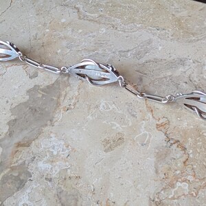 May include: A silver bracelet featuring three stylized floral or leaf-shaped links. Each link has an openwork design and appears to hold a pearl or a similar white stone. The bracelet is laid out on a light-colored, textured surface.