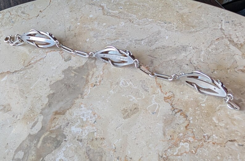 May include: A silver bracelet featuring three stylized floral or leaf-shaped links. Each link has an openwork design and appears to hold a pearl or a similar white stone. The bracelet is laid out on a light-colored, textured surface.