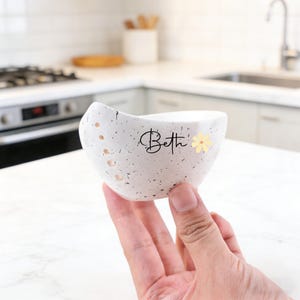 以下が含まれることがあります： 黒い筆記体で「Beth」という名前と小さな黄色の花が描かれた、白い斑点のあるセラミックボウル。ボウルは湾曲した形状で、側面にいくつかの小さな穴があります。ボウルは人の手で持たれています。