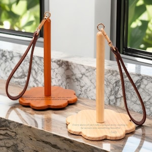 以下が含まれることがあります： 茶色の革ひもが付いた木製ペーパータオルホルダー2つ。ホルダーは花の形をしたベースと木製の棒を持っています。左側のホルダーのベースはオレンジ色で、右側のホルダーのベースは天然木です。ホルダーは、大理石のカウンタートップの上に置かれています。右側のホルダーのベースにあるテキストは、「糸の玉はそれぞれユニークな物語を語り、美しいものへと織り込まれるのを待っています -Judy」と書いてあります