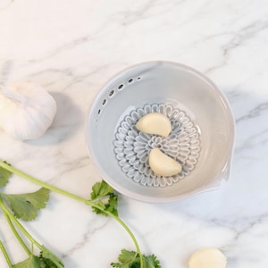 May include: A light gray ceramic garlic grater with a floral design, containing two garlic cloves. A whole garlic bulb and fresh cilantro are also visible. This kitchen tool is designed for grating garlic.