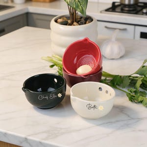 以下が含まれることがあります： 黒、クリーム、バーガンディ、赤の4色のセラミック製ニンニクおろしボウルセット。各ボウルには注ぎ口と小さな花の模様があります。黒いボウルには「Chef Ellie」と書かれています。クリーム色のボウルには「Beth」と書かれています。