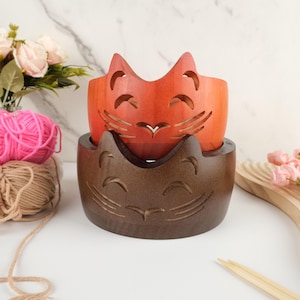 以下が含まれることがあります： 猫の形をした木製の糸巻きボウルが2つ。上のボウルはオレンジ色、下のボウルは茶色。両方のボウルには、笑顔の猫の顔が彫られています。