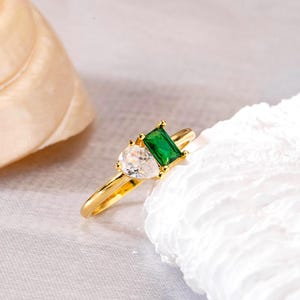 May include: A gold ring featuring a pear-shaped clear stone and a rectangular emerald-cut green stone. The ring is set on a thin gold band. The ring is displayed on a white textured surface.