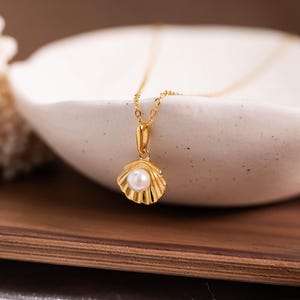 Könnte beinhalten: Eine goldfarbene Halskette mit einem Muschel-Anhänger, der eine kleine, runde Perle enthält. Die Halskette wird in einer weißen Keramikschale präsentiert, die auf einem Holztablett steht. Der Hintergrund zeigt ein strukturiertes, cremefarbenes korallenartiges Objekt.