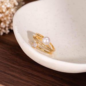 Puede incluir: Un anillo de tono dorado con diseño de doble banda, donde una banda está adornada con una perla blanca y la otra presenta una pequeña piedra transparente de corte redondo junto a una hilera de diminutas piedras brillantes engastadas.