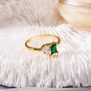 May include: A gold ring featuring a pear-shaped clear stone and a square-cut emerald green stone. The ring is set on a white, textured surface. The ring's band is a thin, polished gold.
