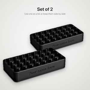 Könnte beinhalten: Ein Set aus zwei schwarzen rechteckigen Schalen mit internen Gitterunterteilungen. Jede Schale zeigt den geprägten Text "Your name here" auf der Vorderseite. Der Text "Set of 2" und "Use one as a lid, or keep them side by side" steht darüber.