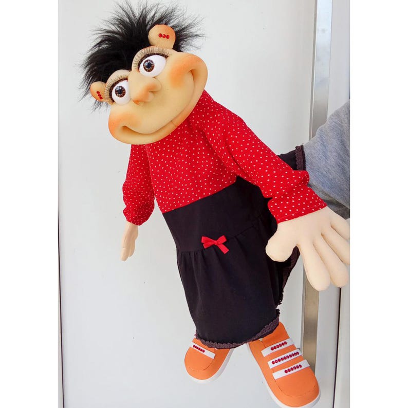 May include: A handmade puppet with a whimsical design. The puppet features a round, pale face with large eyes, a black hair, and a red polka-dot shirt. It wears a black skirt with a red bow and orange shoes with white stripes.