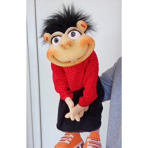 May include: A handmade puppet with a smiling face, black hair, and large eyes. The puppet wears a red polka-dot shirt, a black skirt, and orange sneakers. The puppet's hands are clasped in front of it.