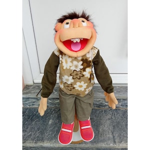 May include: A handmade puppet with a smiling face, brown hair, and a floral patterned shirt. The puppet wears green pants and red shoes. The puppet's mouth is open, revealing white teeth and a pink tongue. The puppet is standing on a wooden stand.