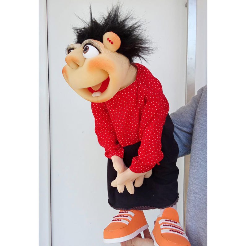 May include: A handmade puppet with a smiling face, black spiky hair, and a red polka dot shirt. The puppet wears a black skirt and orange sneakers. The puppet's hands are clasped together.