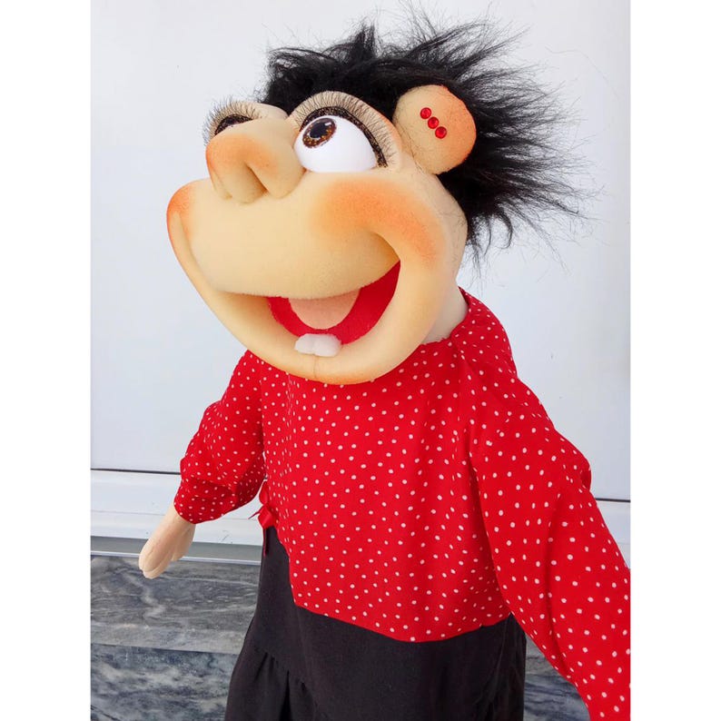 May include: A whimsical puppet with a large, expressive face. The puppet has a tan face with rosy cheeks, large eyes, and a wide, smiling mouth. It wears a red polka-dot shirt and a black skirt. The puppet's hair is black and spiky.