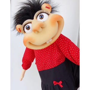 May include: A whimsical doll with a large, expressive face, featuring oversized eyes, a prominent nose, and a wide grin. The doll wears a red polka-dot top and a black dress with a red bow. Its black hair is styled in a wild, spiky fashion.