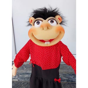 May include: A handmade puppet with a smiling face, black hair, and large blue eyes. The puppet wears a red polka-dot top and a black dress with red bows. The puppet's mouth is open, revealing two white teeth.