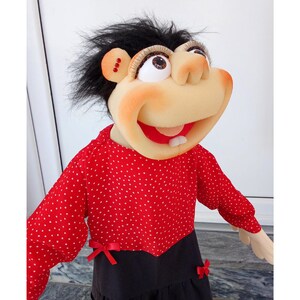 May include: A whimsical puppet with a large, expressive face and black, fluffy hair. The puppet wears a red polka-dot top with a black skirt, accented with red bows. The puppet's features include large eyes, a prominent nose, and a wide, smiling mouth.