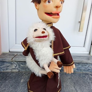 Op de afbeelding: Een handpop van een figuur met bruin haar en een bruine mantel met gouden rand, die een kleinere witte pluche lam-handpop vasthoudt. Het figuur heeft een lichte huidskleur, roze lippen en bruine ogen, terwijl het lam witte vacht en bruine hoeven heeft.