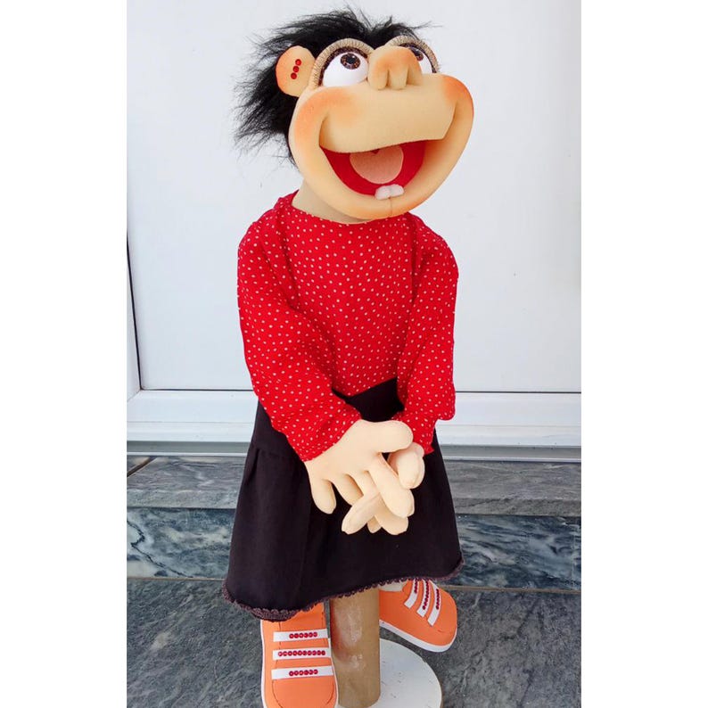 May include: A handmade puppet with a smiling face, black hair, and large eyes. It wears a red polka-dot shirt, a black skirt, and orange sneakers. The puppet's hands are beige, and it stands on a wooden dowel.