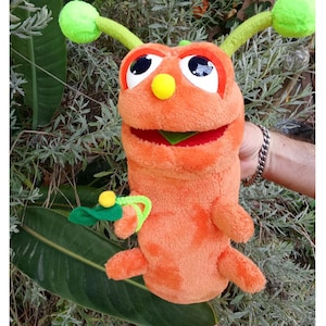 Puede incluir: Títere de mano con forma de oruga naranja con ojos grandes y antenas verdes. El títere tiene una boca roja y un pequeño detalle de hoja verde. Un pequeño accesorio verde y amarillo está unido al costado del títere. Sobre un fondo de follaje verde.