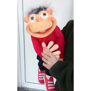 May include: A handmade puppet with a smiling face, freckles, and gray hair. The puppet wears a red long-sleeved shirt, black pants, and red sneakers with white stripes. The puppet's large, expressive eyes and open mouth are visible.