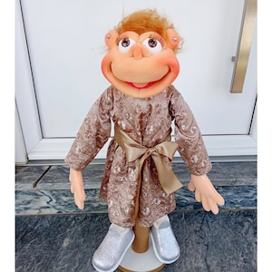 May include: A handcrafted puppet with a monkey-like face, featuring a peach-colored face, large eyes, and a red mouth. It wears a brown floral dress with a gold sash and silver glitter shoes. The puppet has orange hair.