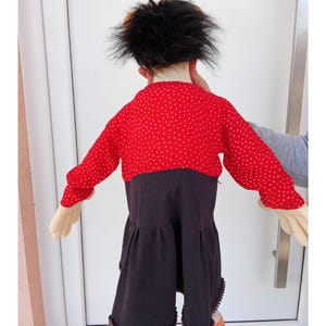 May include: A child's costume featuring a red top with white polka dots and long sleeves, paired with a black skirt. The costume includes a black wig and beige gloves, creating a playful and whimsical look.