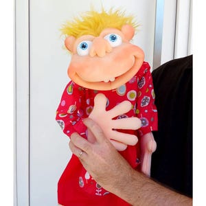Könnte beinhalten: Eine Schaumstoff-Handpuppe mit leuchtend gelben Haaren, großen blauen Augen und einem breiten, lächelnden Mund mit zwei sichtbaren Zähnen. Die Puppe trägt ein rotes Langarmshirt mit einem bunten Muster aus Kreisen und abstrakten Formen.