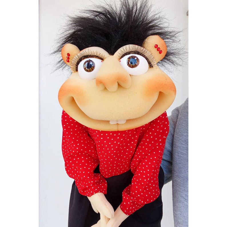 May include: A whimsical puppet with a large, smiling face, prominent nose, and expressive eyes. The puppet has black, spiky hair and wears a red shirt with white polka dots. The puppet's hands are clasped in front of it, and it is set against a white background.