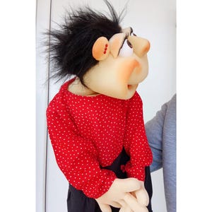 May include: A handmade puppet with a whimsical design. The puppet features a fabric face with a large nose, expressive eyes, and a black, spiky hair. It wears a red polka-dot shirt and black pants. The puppet's hands are visible.