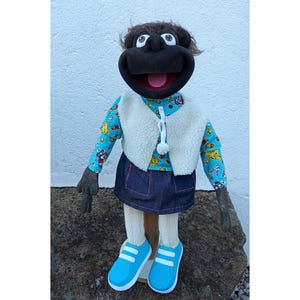 May include: A black hand puppet with brown hair, wearing a blue shirt with a colorful pattern, a white vest, a blue denim skirt, and blue shoes.