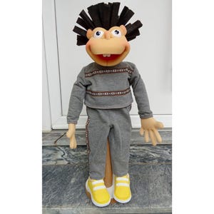 May include: A handmade puppet with a tan face, large eyes, and a wide smile. It wears a gray sweatshirt and sweatpants with decorative trim, and yellow sneakers. The puppet has a dark brown, spiky hairpiece.