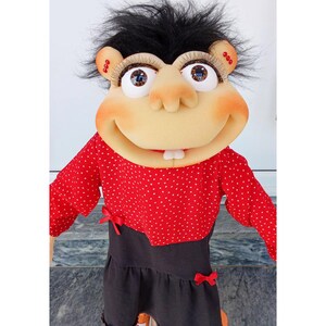 May include: A handmade puppet with a smiling face, large eyes, and black hair. The puppet wears a red polka-dot shirt with red bows and black shorts. The puppet's features include large eyelashes and two front teeth.