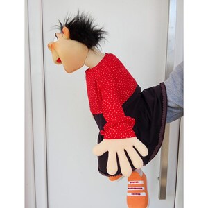 May include: A handmade puppet with a tan face, black hair, and a red polka-dot shirt. The puppet has large, white hands and is wearing an orange shoe. The puppet is leaning forward, as if interacting with something.