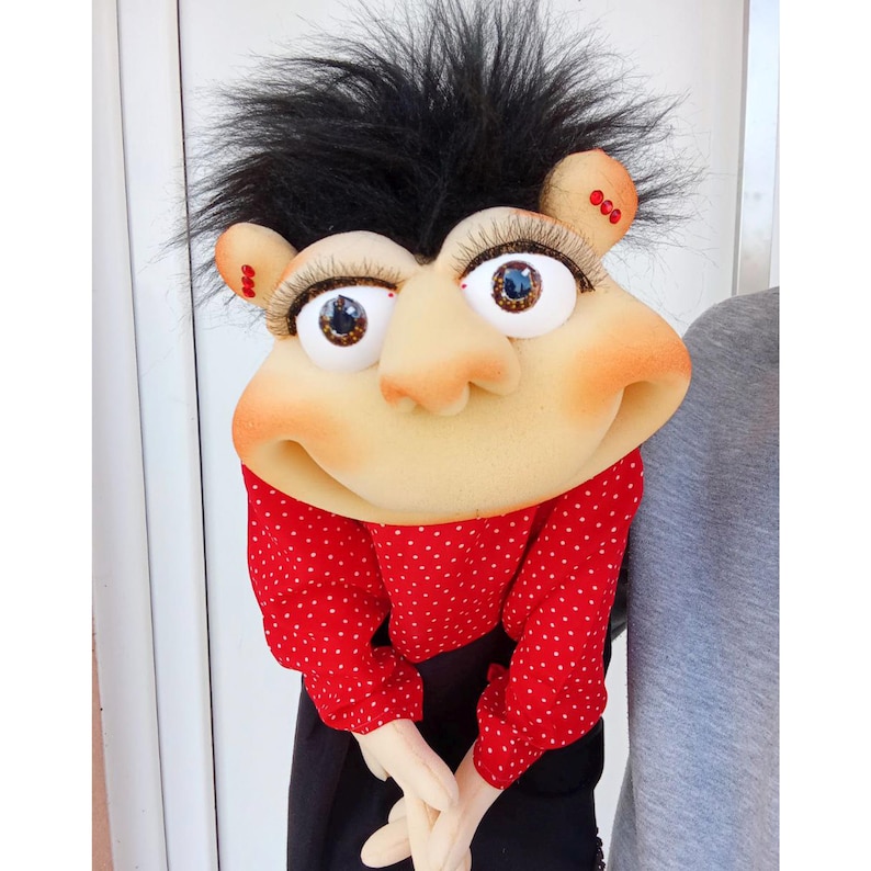May include: A whimsical puppet with a large, expressive face. The puppet has black, spiky hair, large eyes with long eyelashes, and rosy cheeks. It wears a red polka-dot shirt and black pants, with its hands clasped together.