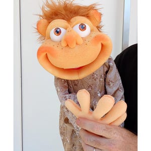 Op de afbeelding: Een schuimrubberen handpop met een oranje huid, warrig oranje haar en grote witte ogen met oranje irissen. De pop heeft een brede, lachende mond, een prominente neus en sproeten. Het draagt een shirt met lange mouwen met een beige en wit bloemenpatroon.