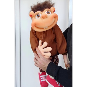 May include: A handmade puppet with a large, smiling face, brown hair, and red accents. The puppet wears a brown shirt, patterned pants, and red shoes with white stripes. The puppet is being held by a person.