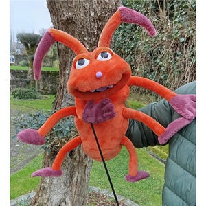 May include: Orange and purple puppet with six legs and a large head. The puppet has a wide mouth with white teeth and a pink nose. The puppet is made of soft, plush fabric.