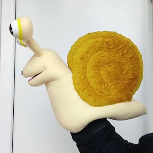 May include: A plush snail puppet with a cream-colored body and a textured, golden-yellow shell. The snail has large, expressive eyes and a smiling mouth. The puppet is designed to be worn on the hand, with the arm acting as the snail's body.