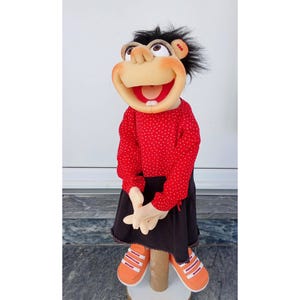May include: A handmade puppet with a smiling face, wearing a red polka-dot shirt, black skirt, and orange sneakers. The puppet has black hair and is posed on a wooden stand. The puppet's mouth is open, revealing a red interior.