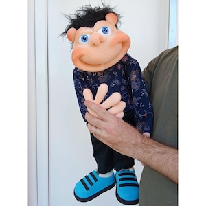 Pode incluir: Fantoche feito à mão com um rosto sorridente, olhos azuis e cabelo preto. O fantoche usa uma camisa azul marinho com um padrão floral, calças pretas e sapatos azuis com listras pretas. O fantoche está sendo segurado por uma pessoa.