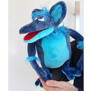 May include: A plush puppet in shades of blue, featuring a long snout, large ears, and a red-lined mouth. The puppet has a light blue chest and accents, with fluffy details around the neck and wrists. The puppet is designed to be held and operated by hand.