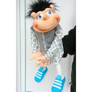 May include: A handmade puppet with a tan face, blue eyes, and black hair. It wears a gray button-up vest over a floral print shirt and blue sneakers with white stripes. The puppet's arms and legs are visible.