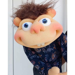 May include: A whimsical puppet with a tan face, large blue eyes, and a pink nose and cheeks. The puppet has brown, messy hair and is wearing a dark blue shirt with a floral pattern. The puppet's mouth is open, revealing two teeth.